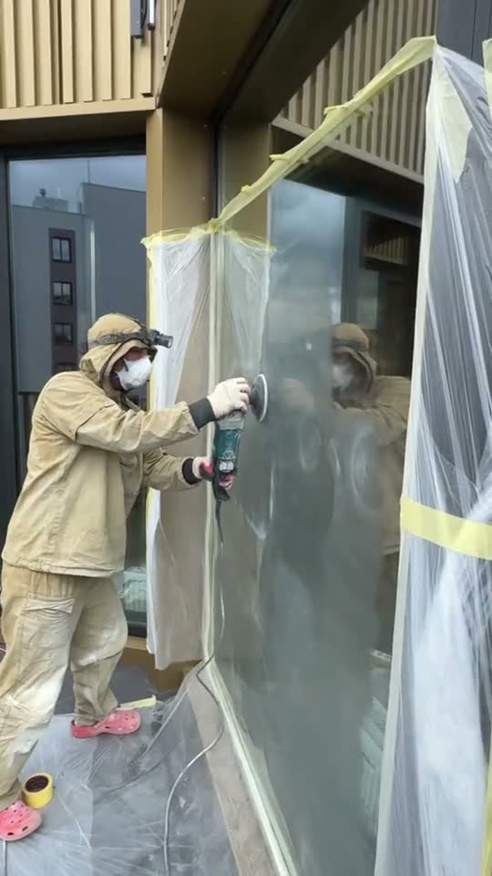 Мастер в защитном костюме полирует фасадное стекло, рама закрыта плёнкой